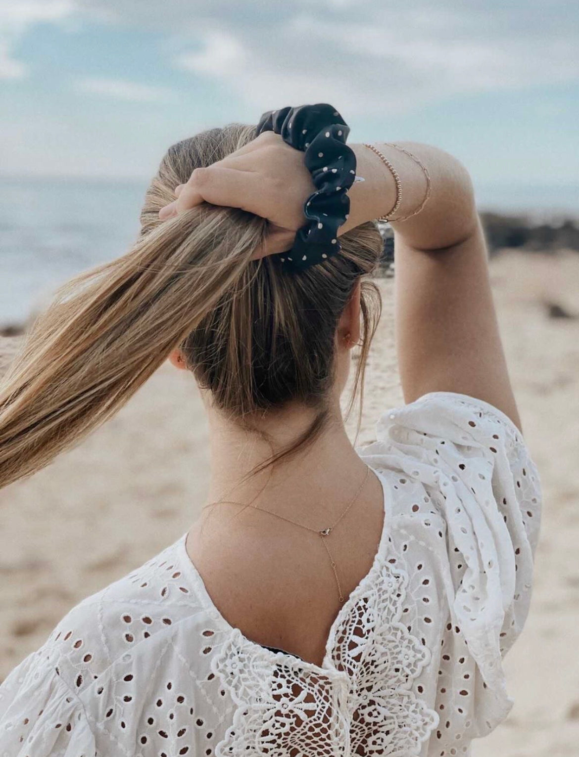 Person holding up their hair with a dark patterned scrunchie around their wrist at the beach